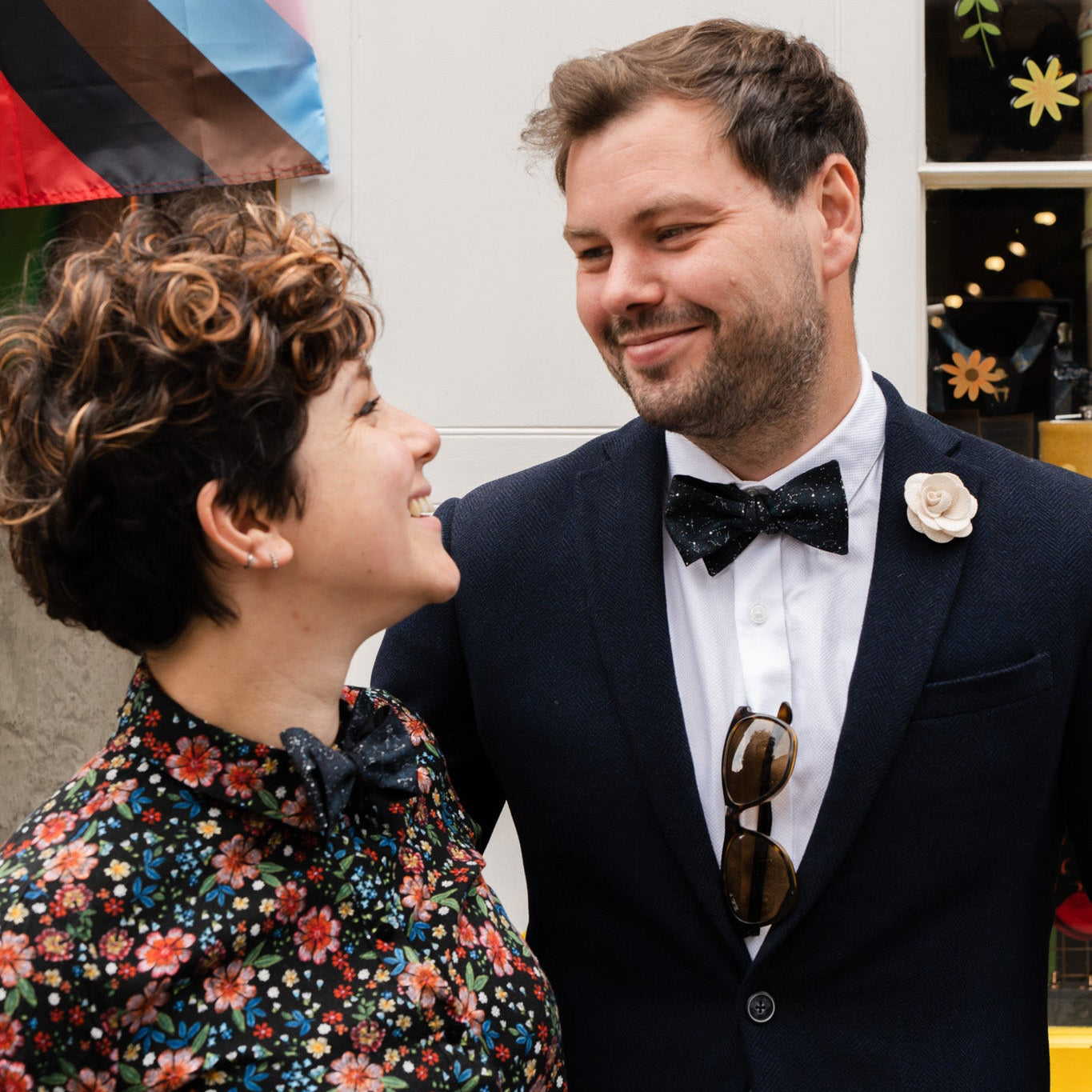 Man in a suit and bow tie standing next to a woman in a floral dress, both looking at each other.